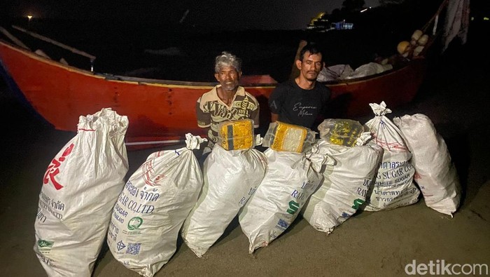 Dua pria terlihat berdiri di samping karung-karung besar yang berisi narkoba yang disita. Penangkapan ini terjadi di malam hari, menunjukkan upaya keras aparat penegak hukum dalam memberantas penyelundupan narkoba.