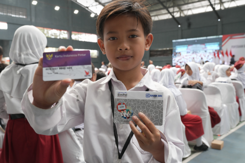 Dalam gambar ini, seorang siswa bangga menunjukkan Kartu Indonesia Pintar (KIP) dan kartu SimPel di sebuah acara resmi. KIP adalah simbol dukungan pemerintah untuk pendidikan yang memastikan anak-anak mendapatkan akses ke bantuan pendidikan, sementara kartu SimPel memudahkan transaksi keuangan.