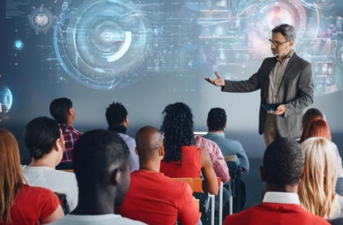  suasana kelas di Ohio State University yang mempresentasikan salah satu momen dari program AI Fluency—seorang dosen sedang membimbing mahasiswa yang tampak fokus di hadapan layar dan perangkat digital